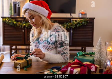Adult female wearing Santa hat fuzzy sweater, sitting at home table wrapping gold gift with ribbon Stock Photo
