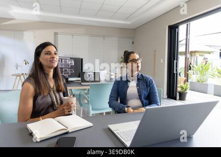 Two adult female colleagues discussing floor plans in modern office with laptop notebook coffee cup Stock Photo