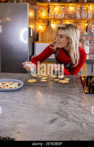 Female baker decorating cookies on rack with piping bag on marble kitchen counter, copy space Stock Photo