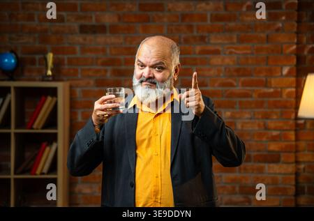 Indian senior businessman or corporate executive drinking and promoting fresh water in a glass while standing against a red brick wall in a cozy offic Stock Photo