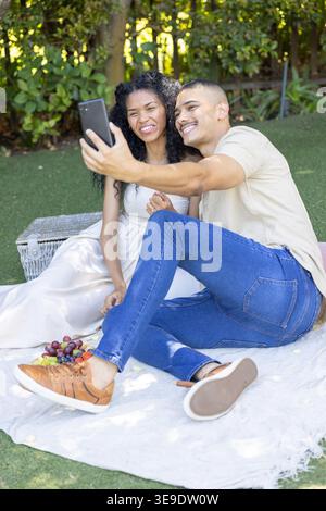 Couple sitting on blanket-in-yard woman in white dress man holding phone near basket and grapes. Selfie, duo, outdoor, picnic, candid, casual, romance Stock Photo