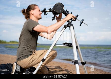 young man prepares his astronomical telescope for use on a sandy beach. astronomer is focused on adjusting equipment under clear blue skies, ready to observe solar activity as part of astronomy hobby Stock Photo
