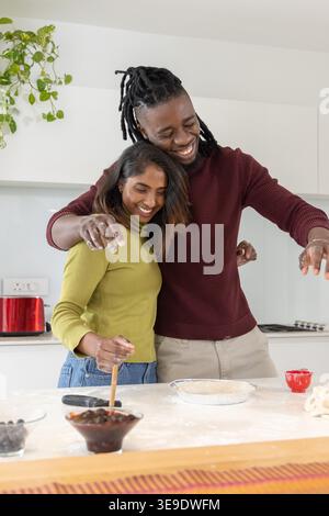Diverse couple embracing and preparing pie in home kitchen with pie tin and wooden spoon. Baking, dough, pastry, crust, flour, berries, cozy Stock Photo