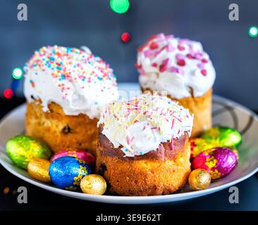 A close-up photograph showcasing three traditional Easter kulich cakes decorated with white glaze and colorful sprinkles. They rest on a table surroun Stock Photo