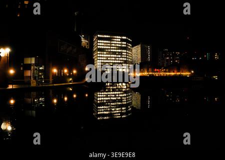 Rows of brightly lit office windows are reflected in the dark waters of a canal at night with a lit pub garden at Gas Street Basin, Birmingham, UK. Stock Photo