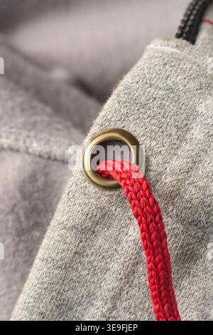 red braided drawstring cord through brass metal eyelet on gray heathered fleece fabric, top view macro closeup, apparel manufacturing fashion detail Stock Photo