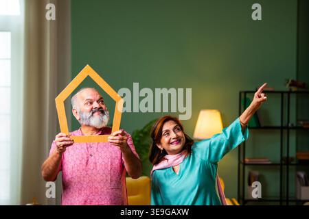 A senior Indian couple in their living room researching real estate. The woman holds a paper home model while the husband uses a laptop to browse prop Stock Photo