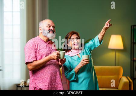 A senior Indian couple stands in their living room holding coffee mugs, looking upward with hopeful expressions. They share a peaceful moment of refle Stock Photo