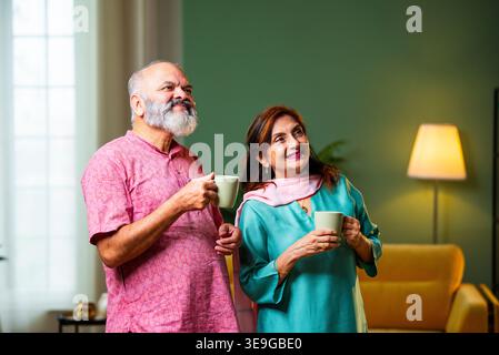 A senior Indian couple stands in their living room holding coffee mugs, looking upward with hopeful expressions. They share a peaceful moment of refle Stock Photo