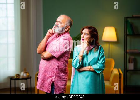 A senior Indian couple standing indoors, lost in deep thought. They use thinking gestures like touching their chins while contemplating a decision or Stock Photo