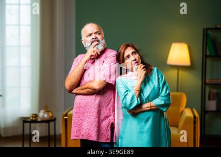 A senior Indian couple standing indoors, lost in deep thought. They use thinking gestures like touching their chins while contemplating a decision or Stock Photo