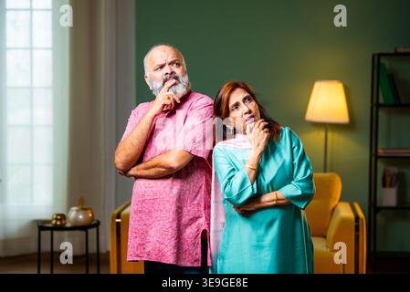A senior Indian couple standing indoors, lost in deep thought. They use thinking gestures like touching their chins while contemplating a decision or Stock Photo