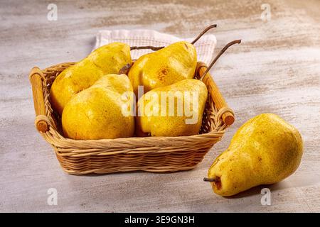 Vibrant yellow pears arranged in rustic wicker basket, one pear rests beside it on light textured wooden surface. Soft natural light creates warm, inv Stock Photo