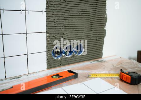Close-up of wall tiling during home renovation. White ceramic tiles on wet adhesive, with electrical boxes exposed. Construction tools like a level tape measure in the foreground. Process concept. Stock Photo