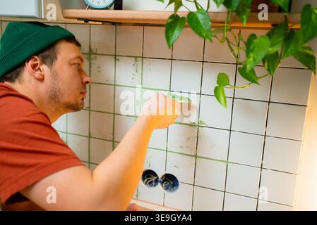 Young man carefully applying green grout to ceramic kitchen backsplash tiles, focused on diy renovation and tiling work, close-up of hands and trowel in modern apartment Stock Photo