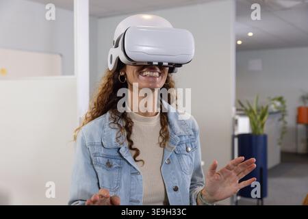 Mature woman wearing white VR headset and denim jacket reaching, using VR content in office Stock Photo