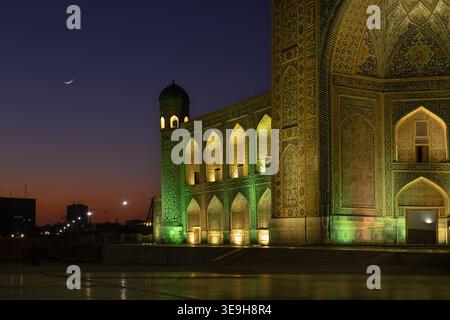 Evening lit facade of Tilya-Kori on Registan square. Evening blue pink sky, young crescent ramadan moon. Large portal arch rich with turquoise mosaic, Stock Photo