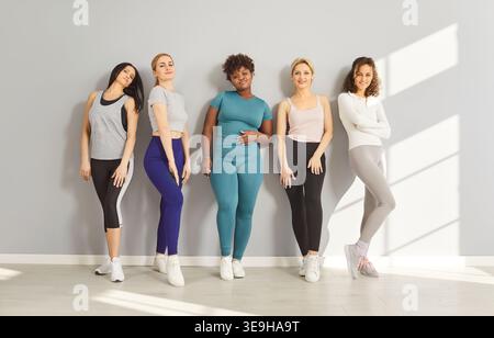 Smiling diverse women in athletic wear posing together against gray wall, showing healthy lifestyle. Stock Photo