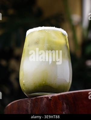 Green drink with salt rim sits on wooden surface. Soft lighting highlights glass and ice. Faded background suggests cozy, relaxed setting. Perfect for Stock Photo