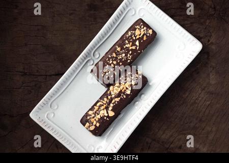 Two chocolate bars sit on a white plate. Nuts sprinkle their surfaces, adding texture. Dark wood background contrasts with the clean plate. Sugary tre Stock Photo