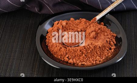 Cocoa powder on a black plate and in a teaspoon, set on a dark bamboo mat. Close up. Stock Photo