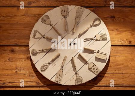 Unique wall clock made from vintage kitchen utensils on a white wooden dial, mounted on a rustic timber wall. Stock Photo