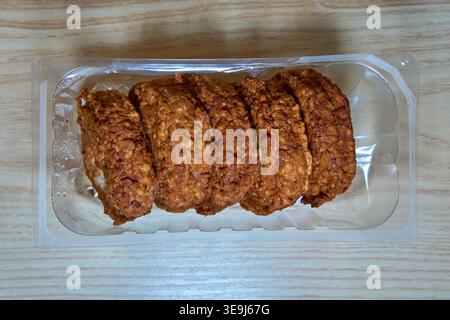 Five golden-brown fried patties in a clear plastic container on a light wooden background. Tasty and convenient ready-to-eat meal. Stock Photo