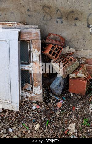 An abandoned weathered wooden window frame leans against a graffiti-covered concrete wall beside broken red bricks and scattered rubble, conveying dec Stock Photo