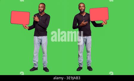 Male model holding red blank speech bubble on camera, creating presentation with empty copyspace cardboard icon. Young adult showing mockup billboard over green screen backdrop. Stock Photo