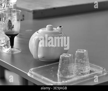 White teapot placed on a tray with two transparent cups, alongside various glassware on a gray table in a dining environment Stock Photo