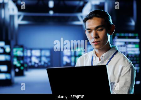 Skilled programmer in data center uses laptop to prevent system overload during peak traffic periods. Experienced professional in server room ensuring enough network bandwidth for smooth operations Stock Photo