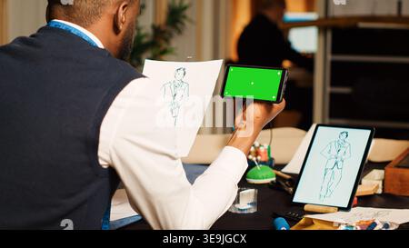 Craftsman using greenscreen in workshop with designers, looking at blank template on mobile phone. Male tailor working with isolated chroma key display, textile manufacturing concept. Stock Photo