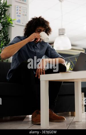 Young arab entrepreneur drinking coffee while sitting on couch in modern business office. Company executive manager enjoying tea while working on laptop in coworking space Stock Photo