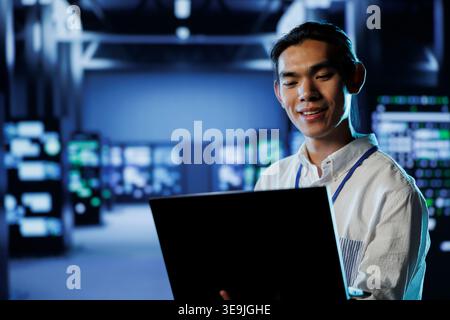 Smiling IT consultant in data center uses laptop to prevent system overload during peak traffic periods. Happy expert in server farm ensuring enough network bandwidth for smooth operations Stock Photo