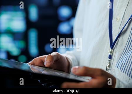Engineer in data center using tablet to prevent system overload, close up. Employee in server room ensuring there is enough network connectivity for smooth operations, blurry background Stock Photo