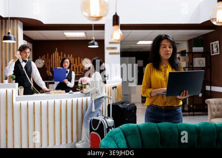 Asian middle aged traveler sitting in reception room holding laptop, walking towards hotel foyer. Woman staying at comfortable resort during holiday carrying notebook in hallway Stock Photo