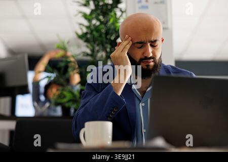 Tired arab businessman with headache working in business office. Overworked executive manager suffering from migraine and rubbing temples while analyzing start up company report on laptop Stock Photo