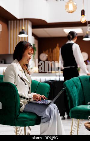 Asian tourist relaxing in welcoming hotel lobby while using laptop. Smiling young traveler enters modern resort and lounges in sitting area before making room reservation Stock Photo