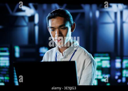 Asian man between server hub rows providing processing resources for businesses worldwide. System administrator fixing data center mainframes tasked with managing massive databases Stock Photo