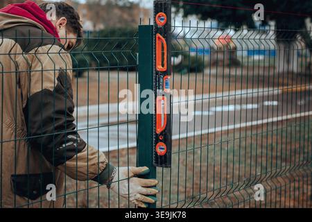 Worker checking vertical alignment of metal fence post using level tool during installation process. Stock Photo