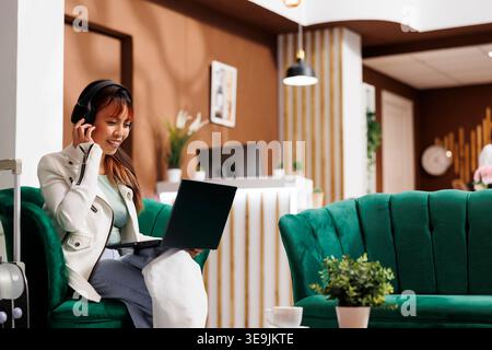 Smiling Asian tourist sitting at laptop with headphones on in modern hotel lounge area, waiting to book room. New customer relaxing in resort lobby before making room reservation Stock Photo