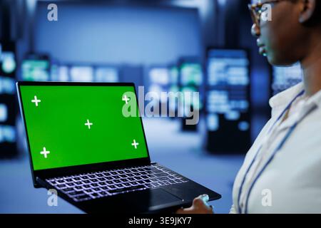 Programmer in server room uses green screen laptop to future proof network from downtimes and unexpected system failures. Tech support with mockup device ensures increased data security Stock Photo