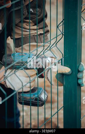 Worker using a power drill to create holes during metal fence installation, close up view of hands and tool in outdoor construction work. Stock Photo