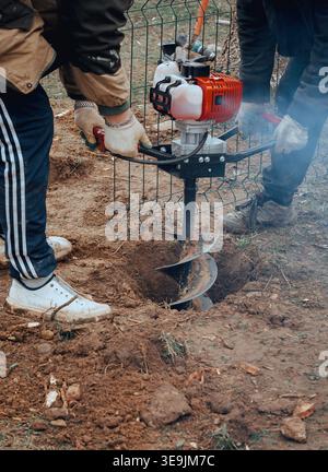 Worker operating a gasoline auger drilling holes in soil, preparation for installation work, outdoor construction process with heavy tool. Stock Photo