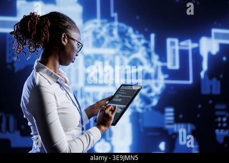 Knowledgeable admin updates neural networks made of interconnected nodes, writing intricate lines of code on tablet. High tech establishment employee uses programming to upgrade AI simulation model Stock Photo