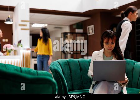 Asian guest getting ready to rent room in welcoming hotel sitting area using laptop. Traveler lounging in modern hotel lobby sitting on sofa working on computer before checking in Stock Photo