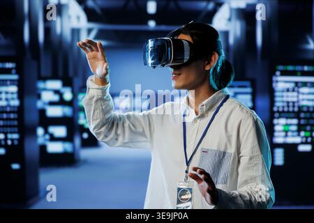Admin in data center uses virtual reality to prevent system overload during peak traffic periods. Man with VR headset in server farm ensuring enough network bandwidth for smooth operations Stock Photo