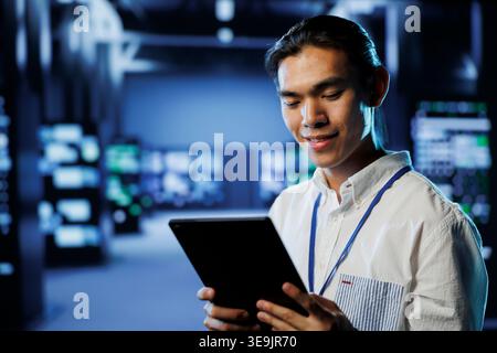 Cheerful computer operator in data center uses tablet to prevent system overload during peak traffic periods. BIPOC man in server facility ensuring enough network bandwidth for smooth operations Stock Photo