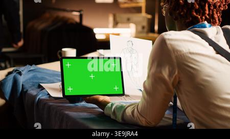 Young couturier looking at greenscreen in tailor workshop, using blank chroma key template at workstation. Female fashion designer working with copyspace display on modern tablet. Stock Photo