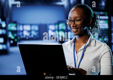 African american woman between server farm rows providing processing resources for businesses worldwide. Admin fixing data center rackmounts tasked with managing massive databases Stock Photo